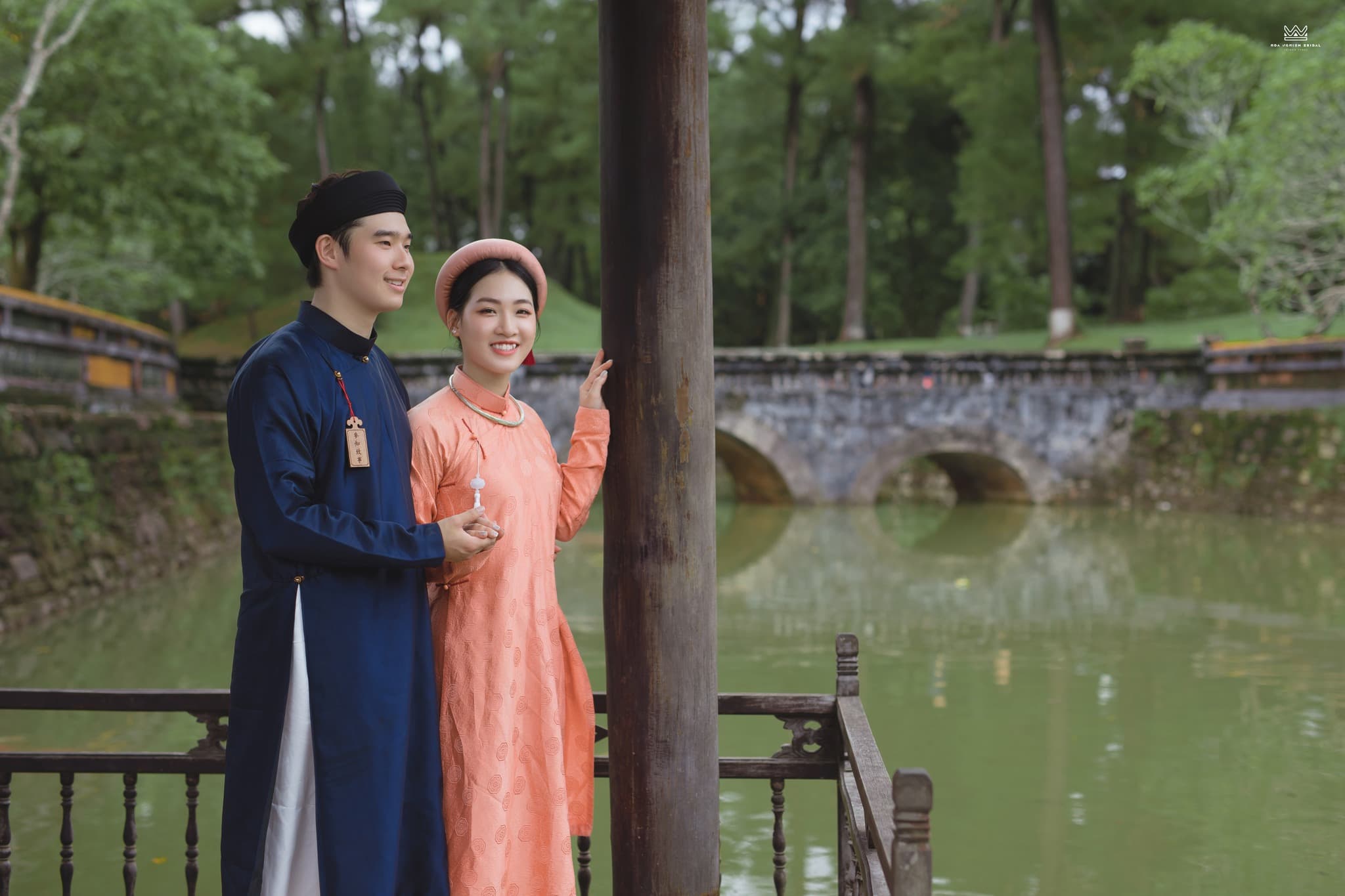 Cổ phục đôi Ngũ Thân Lam Đào Giao Sắc nam nữ đứng cạnh hồ nước trong không gian kiến trúc cổ