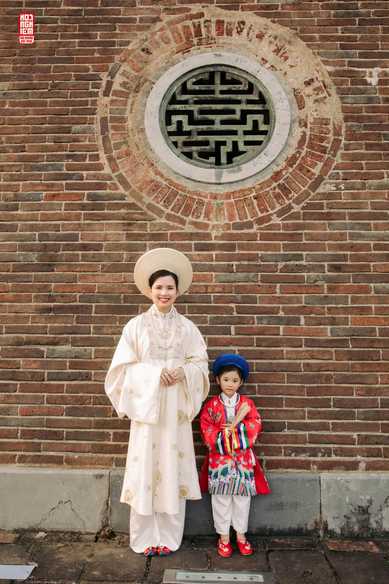 Mẹ và bé gái mặc áo Nhật Bình đứng trước tường gạch cổ với họa tiết truyền thống